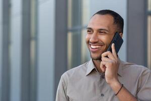 allegro e sorridente africano americano ufficio lavoratore parlando su il Telefono, uomo al di fuori ufficio edificio nel camicia a piedi nel il città, uomo d'affari su pranzo rompere foto