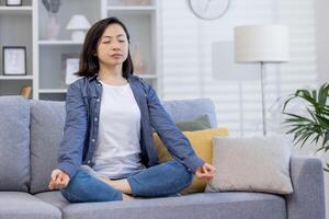 giovane bellissimo asiatico donna solo a casa, Meditare seduta su divano nel vivente camera nel loto posizione, seduta con occhi chiuso a casa. foto