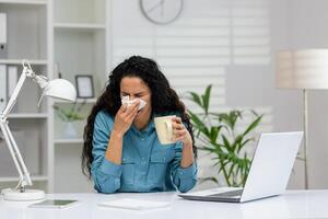 un indisposto femmina dipendente starnuti in un' fazzoletto di carta mentre seduta a sua ufficio scrivania con un' il computer portatile e un' tazza di tè. foto