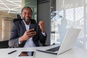 ritratto di riuscito vincitore a posto di lavoro dentro ufficio, africano americano uomo sorridente e guardare a telecamera, uomo d'affari Tenere Telefono, ricevuto in linea notifica di vincente Messaggio. foto