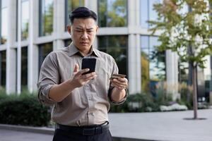 un' preoccupato giovane asiatico uomo è in piedi su un' città strada vicino un' attività commerciale centro, Tenere un' credito carta e guardare a il Telefono nel confusione. foto