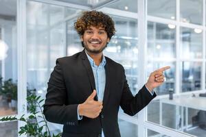 sorridente uomo d'affari dentro ufficio puntamento dita per direzione, contento ispanico capo nel camicia foto