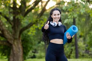 giovane, attivo femmina nel abbigliamento sportivo Tenere un' yoga stuoia e Esprimere a gesti pollici su all'aperto, promozione fitness e salutare stile di vita. foto