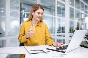 attraente bionda donna lavori nel un' moderno ufficio, si siede a un' scrivania nel davanti di un' computer portatile, detiene un' bicchiere nel sua mano e bevande acqua, il concetto di un' salutare stile di vita, salutare abitudini. foto