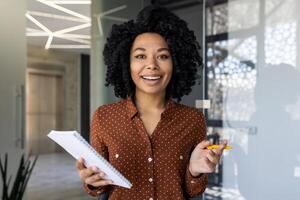 africano americano donna d'affari coinvolgente nel un' chiamata. lei è Tenere documenti e sorridente a il telecamera nel un' ben illuminato, contemporaneo ufficio spazio. foto