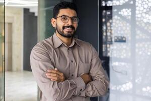 un' professionale giovane uomo con un' barba e occhiali, sorridente con fiducia nel un' ben illuminato, contemporaneo ufficio ambientazione. foto