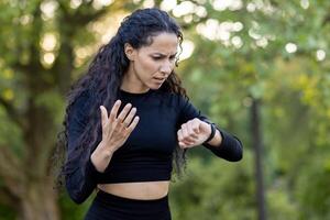 un' confuso ispanico donna nel allenarsi abbigliamento controlli sua orologio durante un esercizio sessione nel un' parco, esprimendo incertezza e leggero frustrazione. foto