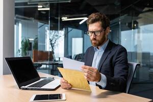 grave e focalizzata maturo capo Lavorando dentro moderno ufficio edificio con computer portatile, uomo d'affari nel attività commerciale completo da uomo accuratamente lettura lettera a partire dal banca, irritato e deluso uomo nel bicchieri e barba. foto