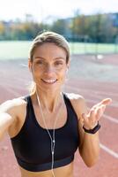 femmina atleta dopo jogging nel stadio parlando con amici e registrazione in linea blog, maturo bionda con cuffie guardare a smartphone telecamera sorridente, chiamata con cuffia. foto