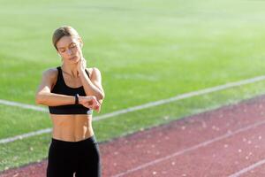 stanco e irritato maturo femmina atleta controllo cuore Vota su inteligente orologio fitness braccialetto, corridore nel stadio dopo attivo esercizio e jogging su soleggiato giorno, bionda donna nel abbigliamento sportivo. foto