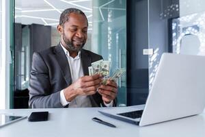 riuscito capo banchiere dentro ufficio seduta a posto di lavoro con computer portatile, contento africano americano investitore Tenere denaro contante i soldi dollari nel mani, uomo conteggio profitto reddito contento con realizzazione risultato. foto