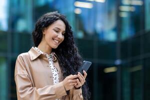 giovane bellissimo ispanico donna a piedi nel il città, attività commerciale donna Tenere Telefono nel mani utilizzando smartphone app, donna sorridente contento e contento al di fuori ufficio edificio con Riccio capelli foto