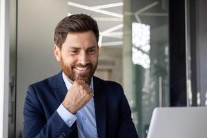 avvicinamento foto di un' giovane uomo d'affari nel un' completo da uomo seduta nel il ufficio nel davanti di un' computer portatile, parlando su un' chiamata, sorridente in il telecamera.