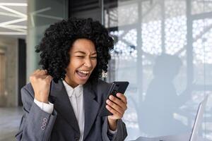 africano americano donna avvicinamento dentro ufficio a posto di lavoro ricevuto in linea Internet notifica vincere Messaggio, attività commerciale donna utilizzando App su smartphone festeggiare trionfo e raggiungimento risultati. foto