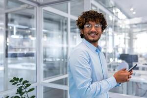 ritratto di riuscito ispanico programmatore ingegnere sviluppatore dentro ufficio, uomo nel camicia e bicchieri Tenere smartphone nel mani sorridente e guardare su finestra, uomo d'affari a posto di lavoro giovane contento. foto