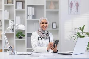 un' allegro femmina assistenza sanitaria professionale nel un' hijab si siede a sua ufficio scrivania, utilizzando un' smartphone e guardare a un' computer portatile, veicolare un' senso di accessibilità e moderno assistenza sanitaria. foto