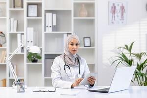 professionale musulmano femmina medico nel un' hijab e bianca cappotto Lavorando a sua ufficio, utilizzando un' tavoletta, con il computer portatile e stetoscopio su scrivania. foto