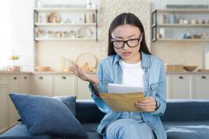 cattivo lettera a partire dal banca, donna seduta a casa deluso e triste lettura Messaggio cattivo notizia, asiatico donna nel cucina foto