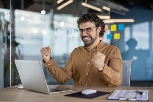 gioioso maschio professionale nel un' casuale camicia giubilante festeggiare un' opera vittoria o successo a il suo ufficio spazio di lavoro con un' il computer portatile. foto