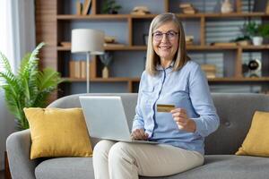 un' donna si siede comodamente su un' divano a casa, shopping in linea utilizzando un' il computer portatile e Tenere un' credito carta, raffigurante convenienza e sollievo. foto