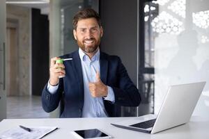 ritratto di un' sorridente giovane uomo seduta nel un' completo da uomo a un' scrivania con un' computer portatile, Tenere un' tosse spray e puntamento super dito a il telecamera. foto