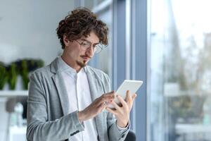 uomo d'affari giovane pensiero grave e concentrato in piedi vicino il finestra dentro il ufficio, il uomo è Tenere un' tavoletta computer nel mani, utilizzando applicazione, appassionatamente lettura a partire dal schermo. foto