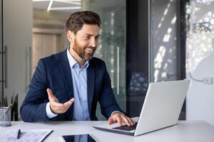 riuscito e allegro giovane uomo d'affari nel un' completo da uomo seduta nel il ufficio a il tavolo con un' il computer portatile e comunicare su un' chiamata , gesticolando con il suo mano. foto