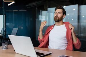 è caldo nel il ufficio, un' uomo d'affari nel un' rosso camicia è piccante, il aria condizionatore è non Lavorando, provando per freddo fuori uso, un' uomo dentro il ufficio è Lavorando con un' il computer portatile. foto