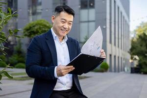 un elegantemente vestito asiatico uomo d'affari recensioni finanziario rapporti con un' lieta Sorridi nel un urbano ambientazione. foto