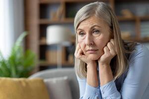 un' di mezza età donna appare preoccupato, seduta solo su un' divano nel un' confortevole casa ambientazione, sua mani su sua testa. foto