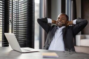 soddisfatto africano americano imprenditore nel un' elegante completo da uomo prende un' tranquillo, calmo rompere a il suo spazio di lavoro con un' il computer portatile. foto