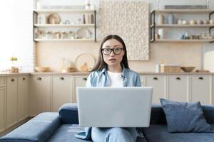 ritratto di un' giovane bellissimo asiatico femmina blogger. seduta a casa su il divano, Tenere un' bianca il computer portatile su il suo giro, guardare intensamente a il telecamera. foto