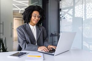 contento e sorridente africano americano donna d'affari digitando su computer portatile, ufficio lavoratore con Riccio capelli e bicchieri contento con realizzazione risultati, a opera dentro ufficio edificio foto