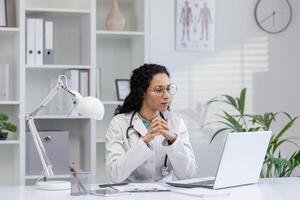 riflessivo ispanico femmina medico seduta a sua scrivania nel un' luminosa medico ufficio, guardare a un' il computer portatile schermo. foto