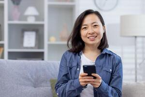 ritratto di un' giovane bellissimo asiatico donna a casa su il divano, donna sorridente e guardare a il telecamera Tenere un' telefono, smartphone applicazione utente, soddisfatto e contento. foto