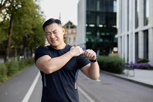 asiatico giovane uomo fare gli sport e in esecuzione su città strada, in piedi e Tenere spalla, sensazione acuto dolore, ferito e slogato. foto