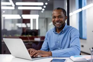 professionale africano americano maschio imprenditore con un' splendente Sorridi a il suo ufficio scrivania utilizzando un' il computer portatile. foto