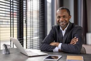 ritratto di un' fiducioso africano americano uomo d'affari con un' caldo Sorridi seduta a il suo scrivania nel un' ben illuminato moderno ufficio ambientazione. foto