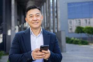 fiducioso asiatico uomo d'affari nel un' completo da uomo sorrisi mentre sms su il suo smartphone, esemplificativo successo e connettività. foto