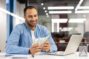 uomo d'affari nel un' blu camicia a il suo scrivania con un' computer portatile, conteggio un' pila di noi dollari, riflettendo finanziario successo e guadagni. foto