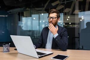 maturo annoiato uomo d'affari Lavorando dentro moderno ufficio costruzione, capo nel bicchieri e attività commerciale completo da uomo utilizzando computer portatile, investitore sbadigli seduta su sedia a tavolo. foto