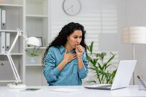 un' professionale donna sensazione male, tosse durante opera nel un' ben illuminato contemporaneo ufficio ambientazione con un' il computer portatile nel davanti di suo. foto