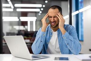 un indiano uomo sensazione stressato e Tenere il suo testa mentre Lavorando su un' il computer portatile nel un' moderno ufficio ambientazione. foto