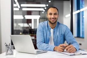 fiducioso indiano uomo con un' amichevole Sorridi Lavorando su un' il computer portatile nel un' ben illuminato contemporaneo ufficio ambientazione. foto