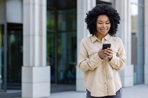 giovane africano americano donna con Telefono sorridente, donna d'affari al di fuori ufficio edificio usi smartphone app, tipi messaggi e sfoglia Internet pagine. foto