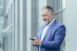 avvicinamento foto. anziano dai capelli grigi uomo, un' uomo d'affari nel un' completo da uomo, sta vicino il ufficio centro e chiamate un' Taxi a partire dal il Telefono, digitando il numero, aspetta, è nel un' fretta per incontrare. foto