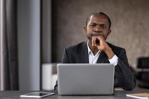 riflessivo adulto maschio nel rigoroso Abiti seduta di portatile computer e sfregamento mento con braccio al chiuso. africano americano persona guardare a telecamera e sensazione confuso con attuale problema soluzione. foto