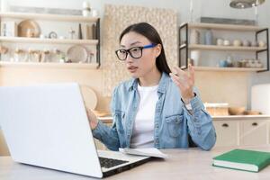 giovane bellissimo donna parlando su chiamata, asiatico donna a casa seduta su divano nel cucina parlando remoto utilizzando il computer portatile per comunicazione e in linea applicazione. foto