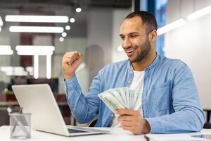 un' contento uomo d'affari a un' scrivania con un' computer portatile, allegramente festeggiare finanziario successo mentre Tenere denaro contante nel mano. foto