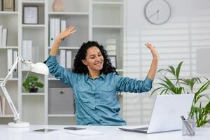 gioioso donna d'affari con braccia sollevato nel eccitazione a sua scrivania nel un' moderno ufficio, festeggiare professionale risultato. foto